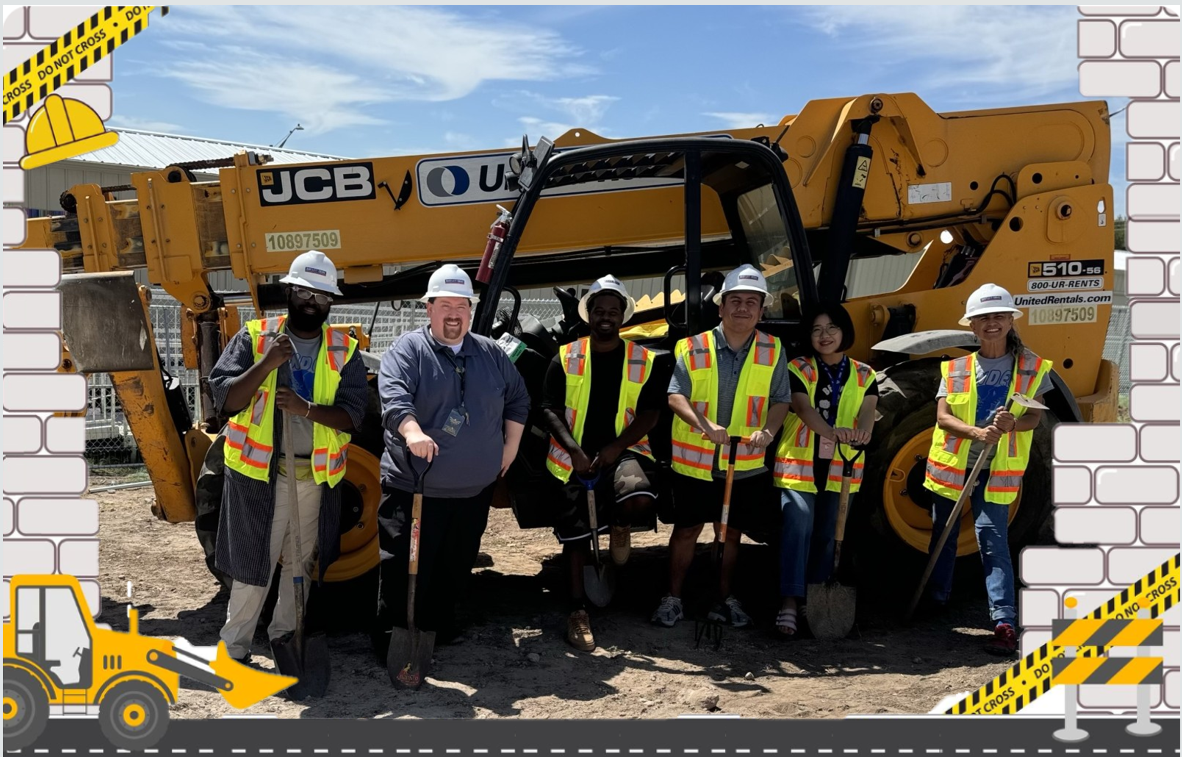 Picture of teachers in a construction site holding shovels in front of heavy-duty construction equipment.